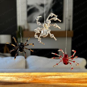 May include: Three hanging spider ornaments in black, clear, and red. The spiders are made of beads and wire, with the text "What can be made with the how-to guide" and "No phycial items are included" at the bottom.