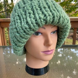 May include: A light green, chunky knit hat. The hat is made of a thick, textured yarn and has a rounded crown and a slightly turned-up brim. The hat is displayed on a mannequin head.