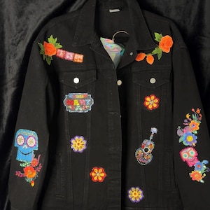 May include: Black denim jacket adorned with colorful embroidered patches. Features include floral and skull designs, a guitar, and the word "LOVE". The jacket has a classic button-up front and two chest pockets.
