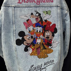 May include: Light blue denim jacket with a rhinestone design featuring Mickey Mouse and friends. The word "Disneyland" is spelled out in pink rhinestones across the top, and "Mickey Mouse" is written in script at the bottom.