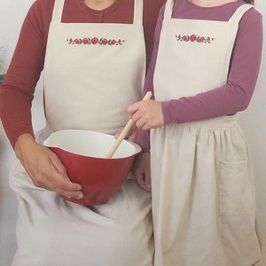 Può includere: Due grembiuli beige con ricamo di rose rosse. I grembiuli hanno spalline regolabili e una grande tasca sul davanti.