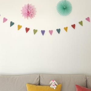 May include: A colorful heart-shaped garland hangs against a white wall, with a pink and a teal paper decoration. A plush bunny sits on a yellow pillow, with a red pillow next to it. The garland is made of felt hearts in various colors.
