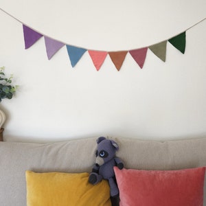 Könnte beinhalten: Eine bunte Stoffgirlande mit dreieckigen Flaggen in den Farben Lila, Rosa, Rot und Grün hängt über einem grauen Sofa mit zwei Kissen. Ein plüschiges graues Stofftier sitzt auf dem Sofa.
