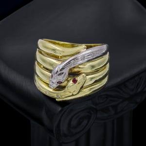 May include: A multi-coil ring featuring a gold band shaped like a coiled snake. The head of the snake is crafted in silver-toned metal with small embedded clear stones and two red stones for eyes, while the body wraps around the finger multiple times.