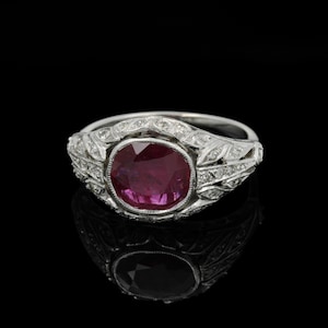 May include: A vintage ring featuring a large, oval-cut ruby gemstone. The ring is made from a silver-coloured metal, with detailed leaf and diamond-shaped patterns along the band. The ruby has a rich, deep red colour.