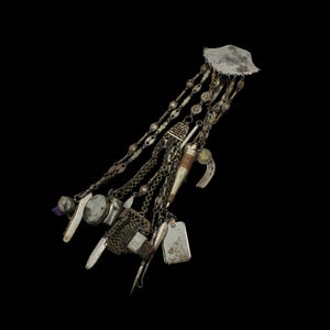 Può includere: Un'antica collezione di ciondoli in argento con vari pendenti, tra cui un piccolo ferro di cavallo, una targhetta identificativa e un coltello in miniatura, sospesi a delicate catene. La collezione è sormontata da un pezzo di metallo decorativo smerlato. Lo sfondo è nero.