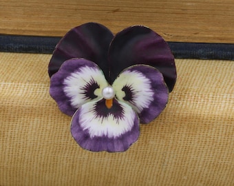 以下が含まれることがあります： パールをあしらった紫と白のエナメル製のパンジーの花のブローチ。ブローチは茶色の布地に置かれています。
