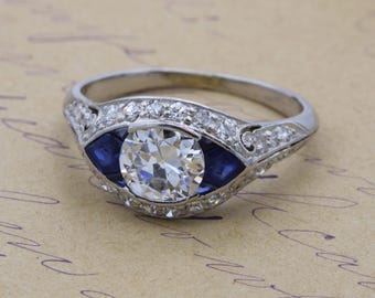 May include: A silver ring featuring a large, round, clear diamond center stone, flanked by two blue stones. The band is adorned with smaller, clear stones. The ring is set against a light brown background with script.