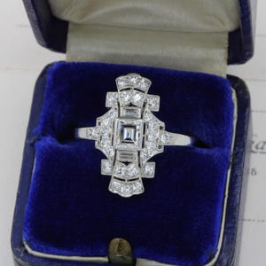 May include: A silver Art Deco ring featuring a central square diamond and surrounding smaller diamonds. The ring is set in a geometric pattern and presented in a blue velvet box. The design includes baguette and round-cut diamonds, showcasing a symmetrical arrangement.