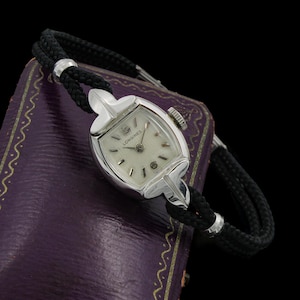 May include: A silver-toned Longines wristwatch featuring a textured white dial with silver hour markers and hands. The watch case has a unique geometric shape and is attached to a black braided cord strap with silver-toned metal accents.