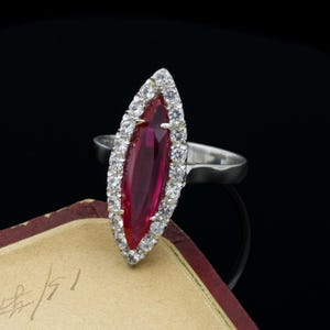 May include: A silver ring featuring a marquise-cut, deep red gemstone, surrounded by a halo of small, clear stones. The ring's design is elongated, with the central stone catching the light. The ring is set against a dark background.