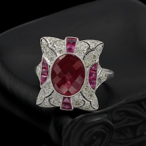 Puede incluir: Un anillo de plata vintage con un gran rubí ovalado facetado en el centro. El rubí está rodeado de rubíes más pequeños y piedras de acento claras, en un diseño ornamentado de estilo Art Déco. El anillo se muestra sobre un fondo oscuro.