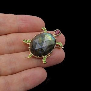 May include: A turtle-shaped brooch crafted in gold, featuring a large, faceted, iridescent labradorite shell. The head is set with a smooth, teardrop-shaped red gemstone, while the four legs are accented with small, bright green peridot stones.
