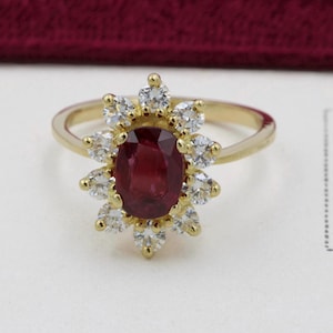 Peut inclure: Une bague en or avec une pierre centrale ovale rouge, entourée d'un halo de petites pierres précieuses rondes et claires. Le corps de la bague est en or uni. Les pierres sont disposées en motif floral ou en forme de soleil.