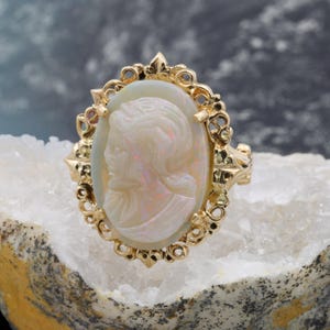 May include: A gold ring featuring an oval cameo with a profile of a woman. The cameo is set in a decorative gold frame. The ring is displayed on a white, crystalline surface against a blurred background.