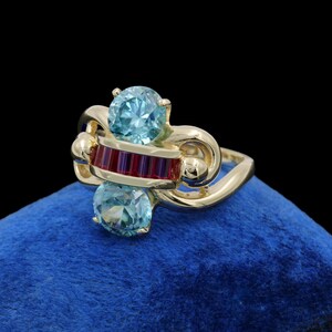 May include: A gold ring featuring two round, light blue faceted stones positioned vertically. Between the two blue stones is a horizontal bar set with five small, rectangular, deep red stones. The gold band has a smooth, polished finish.