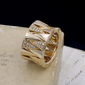 Puede incluir: Un anillo dorado con un diseño geométrico. El anillo presenta secciones triangulares con pequeñas piedras claras incrustadas. El anillo está sobre una superficie clara con texto.