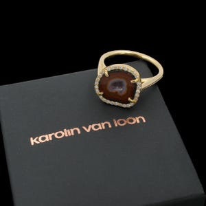 Puede incluir: Un anillo dorado con una piedra central cuadrada marrón, rodeada de pequeñas piedras transparentes. El anillo está sobre una caja negra con el nombre "karolin van loon" en texto color cobre.