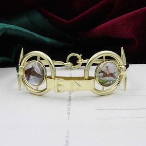 May include: A gold-toned bracelet featuring two circular elements with painted miniature portraits of horses and riders. The bracelet has a central strap with a buckle and is set against a dark background.