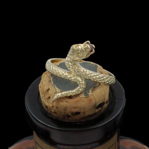May include: A gold snake ring with a red gemstone eye is coiled around a cork. The snake's body has a textured, scale-like pattern. The ring is set against a dark background, highlighting the jewelry's details.