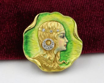 May include: A vintage gold-toned brooch featuring a woman's profile with blonde hair, a jeweled headband, and a diamond earring. The woman's profile is set against a green and yellow enamel background, with a gold border. The brooch is set on a burgundy velvet surface.