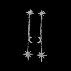 May include: A pair of silver-toned drop earrings featuring a star-shaped stud at the top, connected by a delicate chain to a crescent moon charm and a larger star-shaped pendant at the bottom, all encrusted with small clear sparkling stones.