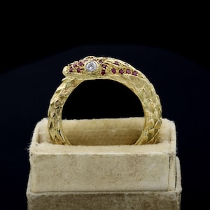 May include: A gold-colored snake ring with a textured surface. The snake's head features a central diamond and smaller red gemstones. The ring is presented in a beige jewelry box, set against a dark background.