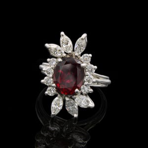 May include: A stunning ring featuring a large, oval-cut, deep red gemstone surrounded by a halo of smaller, round diamonds and marquise-cut diamonds. The ring's band is a bright silver color, and the setting is intricate and detailed. The ring is displayed against a black background.