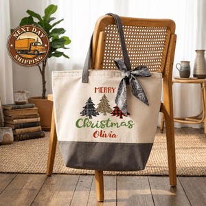 A beige tote bag with a Christmas tree design and the text "Merry Christmas Olivia" printed on it, hanging from a wooden chair.