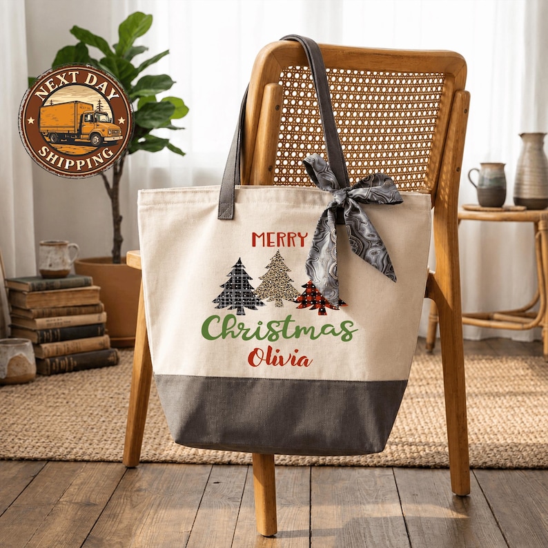 A beige tote bag with a Christmas tree design and the text "Merry Christmas Olivia" printed on it, hanging from a wooden chair.