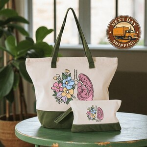 A white tote bag with a colorful floral design and a smaller bag with a similar floral design are displayed on a green table.