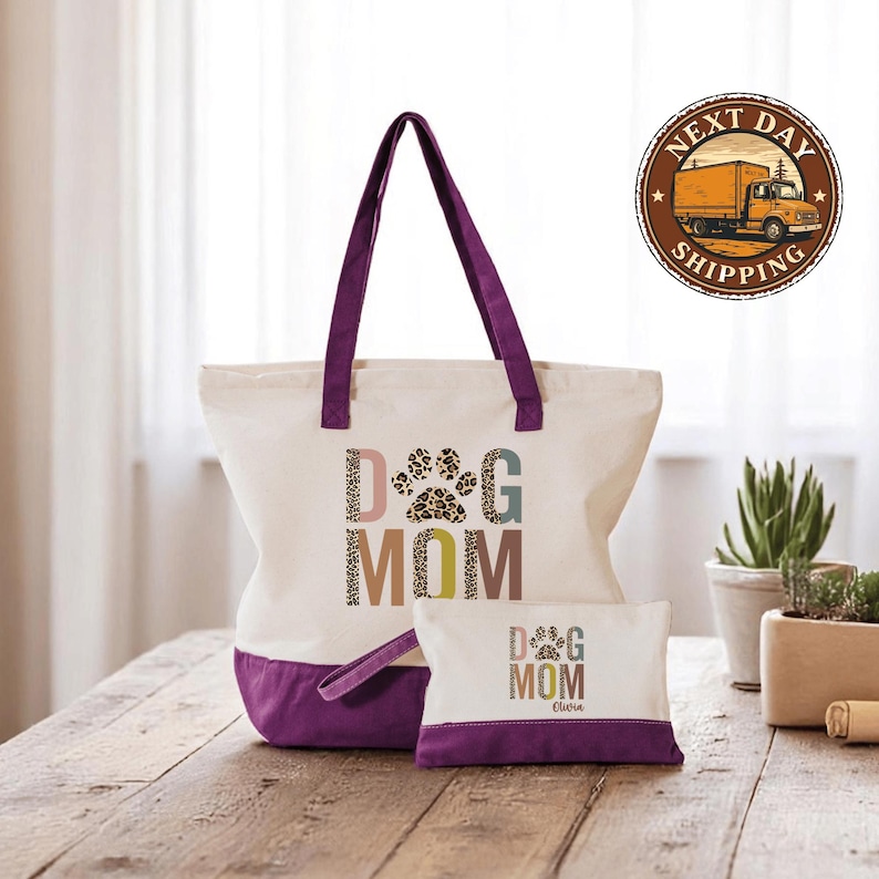 A white tote bag with a purple handle and a smaller bag with a purple handle, both featuring the text "DOG MOM" printed on them, placed on a wooden surface.