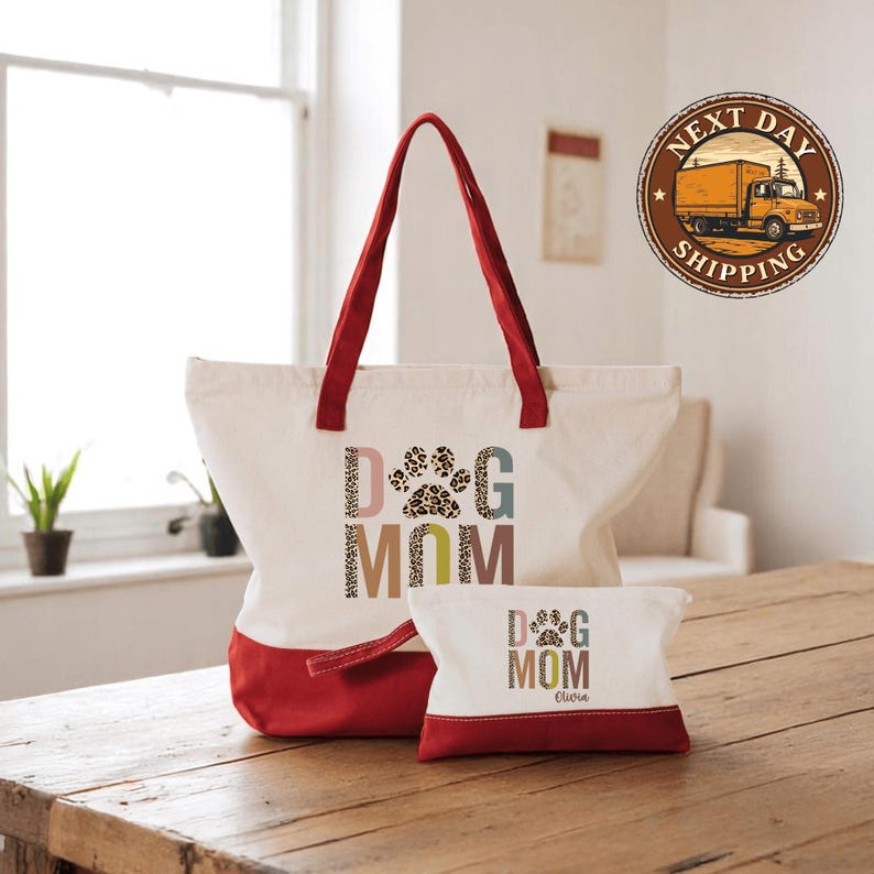 A large tote bag with a red handle and a smaller dog mom bag are placed on a wooden table, with a window and a truck logo visible in the background.