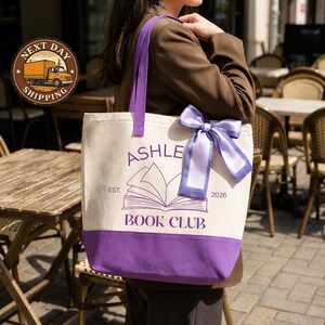 A person is standing in front of a table with chairs, wearing a white tote bag with purple trim and a purple ribbon.