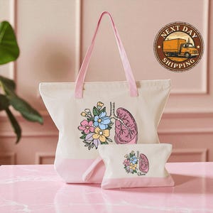 A white tote bag with a pink handle and a smaller white bag with a pink handle, both featuring floral designs, are placed on a pink marble surface.