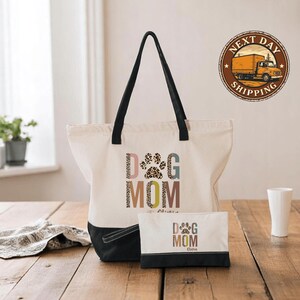 A white tote bag with the text "DOG MOM" printed on it, along with a smaller bag and a white cup on a wooden surface.