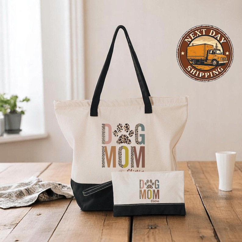 A white tote bag with the text "DOG MOM" printed on it, along with a smaller bag and a white cup on a wooden surface.