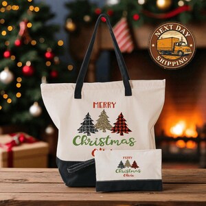 A white tote bag with a Christmas tree design and the text "Merry Christmas" printed on it, along with a smaller bag with the same design and text, placed on a wooden surface in front of a Christmas tree and a fireplace.