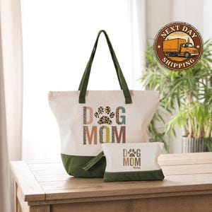 A white tote bag with the text "DOG MOM" printed on it, along with a smaller bag that says "DOG MOM" on it, placed on a wooden surface. There is also a "NEXT DAY SHIPPING" sticker visible in the background.