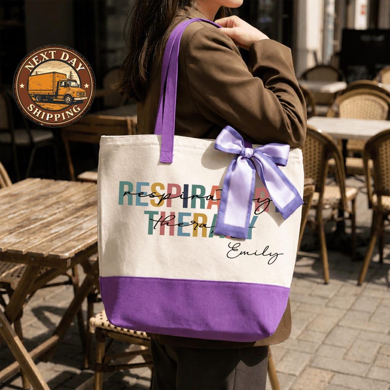 A woman is standing in front of a table with chairs, wearing a tote bag with a purple ribbon and text printed on it.