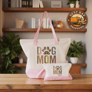 A large tote bag with the text "DOG MOM" printed on it, and a smaller bag with the same text, both sitting on a wooden surface. In the background, there are shelves with various items, including books and plants.