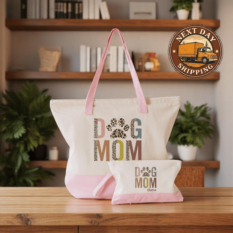 A large tote bag with the text "DOG MOM" printed on it, and a smaller bag with the same text, both sitting on a wooden surface. In the background, there are shelves with various items, including books and plants.