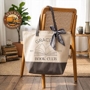 a gray and white tote bag with the text "Grace Book Club" printed on it, hanging on a wooden chair. The bag is placed on a wooden floor, and there are several books and decorative items visible in the background.