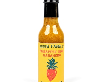 Könnte beinhalten: Eine klare Glasflasche mit scharfer Soße und schwarzem Verschluss. Das gelbe Etikett trägt die Aufschrift "REES FAMILY PINEAPPLE LIME HABANERO HOT SAUCE" mit einem roten Erdbeer- und grünen Ananas-Design. Die Soße im Inneren hat eine goldene Farbe.