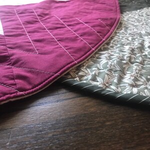 Peut inclure: Deux cercles de tissu matelass&eacute;s, l'un bordeaux avec des coutures blanches et l'autre vert avec un motif floral blanc. Les cercles de tissu sont pos&eacute;s sur une surface en bois fonc&eacute;.