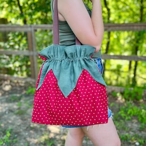 May include: A red tote bag with white polka dots, designed to resemble a strawberry. The bag features a green leaf-shaped top and brown straps. The bag is made of fabric and is suitable for carrying items.
