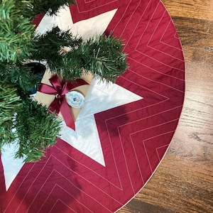 Peut inclure: Une jupe de sapin de No&euml;l matelass&eacute;e rouge et blanche avec un motif en chevron. La jupe est d&eacute;cor&eacute;e d'une &eacute;toile blanche au centre.
