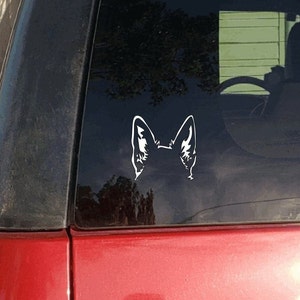 May include: White vinyl decal of a dog's ears, applied to a car window. The decal is a silhouette of the ears, with detailed inner ear markings. The car is red with a black trim.