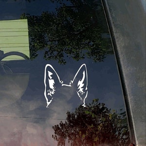 May include: A white vinyl decal of a dog's ears and the top of its head, affixed to a car window. The design is a stylized silhouette, with detailed inner ear markings. The background shows a reflection of trees and sky.