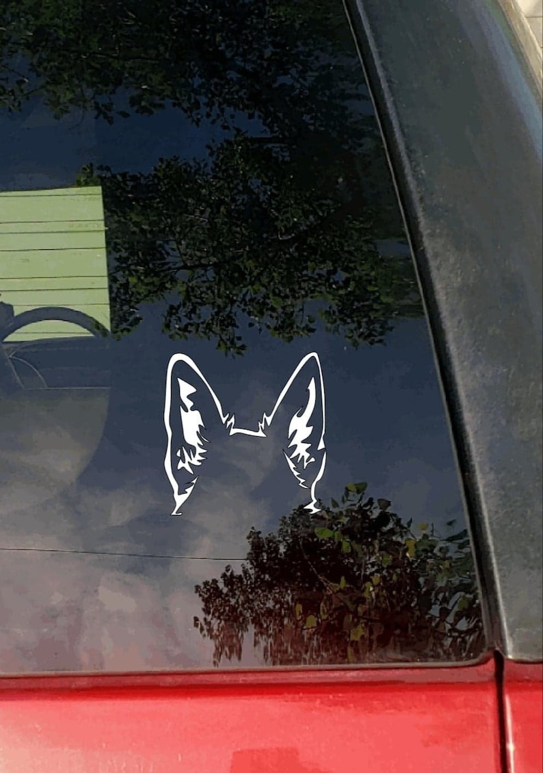May include: A white vinyl decal of a dog's ears and the top of its head, affixed to a car window. The design is a stylized silhouette, with detailed inner ear markings. The background shows a reflection of trees and sky.
